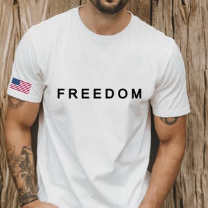 May include: White t-shirt with the word "FREEDOM" printed in black across the chest. An American flag is on the left sleeve. The shirt is made of a soft material and is suitable for casual wear.
