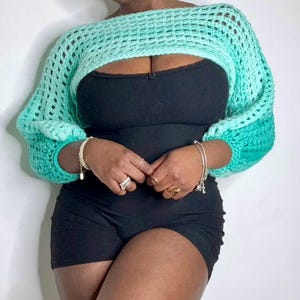 May include: A light green crochet top with a wide, open neckline and long sleeves. The top is made with a loose, open stitch and has a gradient of color from light green to dark green.
