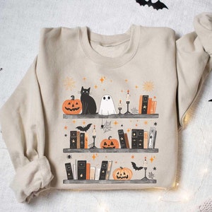 May include: A beige sweatshirt with a Halloween-themed design featuring a black cat, a ghost, pumpkins, and books on a shelf. The design is surrounded by stars and bats.