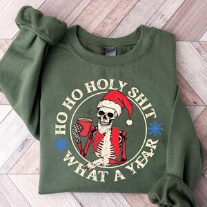 May include: Green sweatshirt with a white circle graphic featuring a skeleton wearing a Santa hat and holding a cup of coffee. The text around the circle reads "Ho Ho Holy Shit What A Year".
