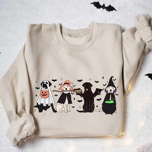 May include: A beige sweatshirt with a graphic print of four dogs dressed in Halloween costumes. The dogs are wearing a ghost costume, a witch costume, a vampire costume, and a witch costume with a cauldron. The graphic is surrounded by bats and stars.