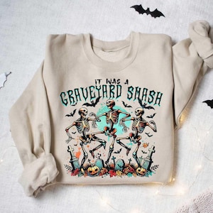 May include: A beige sweatshirt with a graphic print of three skeletons dancing in a graveyard. The text "It was a Graveyard Smash" is printed above the skeletons.