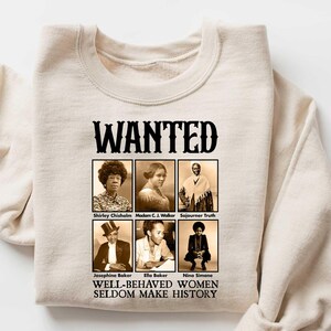 May include: A beige sweatshirt with a black graphic print that says "WANTED" and features a collage of black and white photos of historical figures. The text below the photos reads "WELL-BEHAVED WOMEN SELDOM MAKE HISTORY."