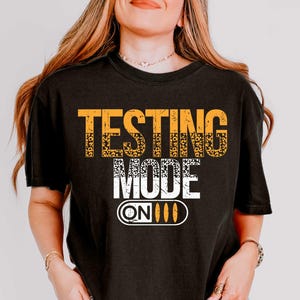 May include: Black t-shirt with the text "Testing Mode On" in yellow and white lettering. The text is stylized to look like a switch with the word "On" and three lines.