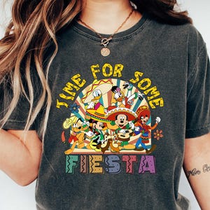 Pode incluir: Camiseta cinza escuro com um gr&aacute;fico colorido com personagens da Disney em trajes festivos. O texto "TIME FOR SOME FIESTA" &eacute; exibido acima e abaixo dos personagens. A camiseta &eacute; combinada com jeans azuis e t&ecirc;nis brancos.