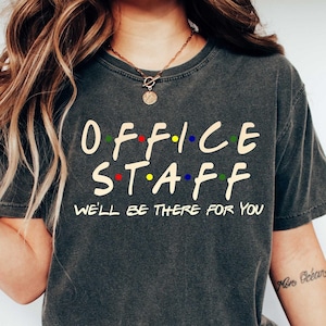 May include: A dark gray t-shirt with the text "OFFICE STAFF" in a stylized font with colorful dots. The text "WE'LL BE THERE FOR YOU" is printed below the main text.