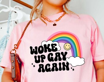 May include: A light pink t-shirt with the text "WOKE UP GAY AGAIN" in black, with a rainbow, smiley face, and clouds graphic. The shirt is a casual style, suitable for everyday wear.
