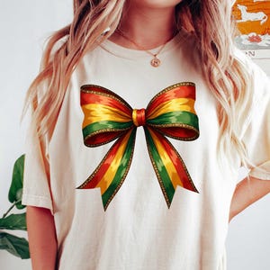 May include: A white t-shirt with a large, colorful bow printed on the front. The bow is made up of red, yellow, and green stripes.