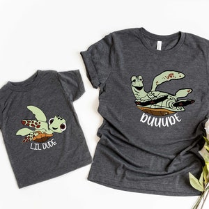 May include: Two gray t-shirts with a cartoon sea turtle design. The larger shirt says "DUDE" and the smaller shirt says "LIL DUDE".