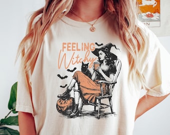 Könnte beinhalten: Ein beiges T-Shirt mit einem Aufdruck einer Hexe, die auf einem Stuhl mit einer schwarzen Katze und einem Kürbis sitzt. Der Text "Feeling Witchy" ist auf dem Shirt aufgedruckt.