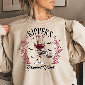 May include: A beige sweatshirt with a graphic design of a skeleton hand holding a glass of red wine. The design includes the text "Rippers Cocktail Club" and a wreath of leaves with bats.