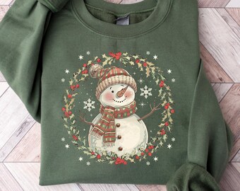 May include: Green crewneck sweatshirt featuring a snowman design. The snowman wears a hat and scarf, surrounded by a wreath of holly and snowflakes. The sweatshirt is a solid green color, perfect for the holiday season.