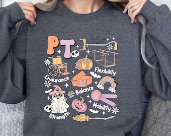 May include: Dark gray sweatshirt with a colorful graphic design featuring the letters "PT" and illustrations related to physical therapy, including a wheelchair, pumpkin, and ghost. The design also includes the words "Endurance", "Strength", "Balance", "Flexibility", and "Mobility".