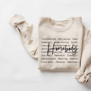 Może przedstawiać: Beżowa bluza z napisem "Homebody" napisanym czarnym, ozdobnym pismem. Bluza zawiera listę sł&oacute;w opisujących osobowość "domatorka", takich jak "introwertyk", "samotnik", "przytulny", "domator", "cichy", "spokojny", "pacyficzny", "spokojny", "wygodny", "skromny", "rozluźniony", "zamyślony" i "nawykowy".