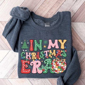 Op de afbeelding: Een donkergrijs sweatshirt met de tekst "In My Christmas Era" in kleurrijke, gestileerde letters. Naast de tekst is een discobal-afbeelding te zien.