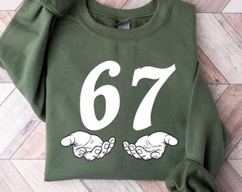 May include: An olive green sweatshirt with the number "67" in white, along with two white, open hands below the number. The sweatshirt is folded on a light brown and white wooden surface.