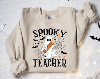 Może przedstawiać: Beżowa bluza z nadrukiem przedstawiającym nauczyciela w duchu Halloween. Nadruk przedstawia ducha trzymającego ołówek i pajęczynę. Tekst "Spooky Teacher" jest nadrukowany czarnym kolorem.