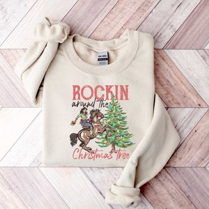 May include: A beige sweatshirt with the text "Rockin' around the Christmas Tree" and a graphic of a person riding a horse in front of a Christmas tree.
