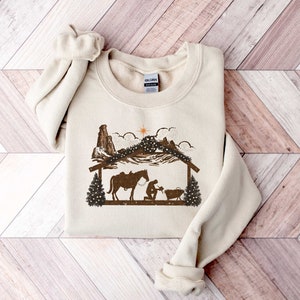 Puede incluir: Sudadera de color crema con un gr&aacute;fico marr&oacute;n de una escena de la natividad con un caballo, una figura arrodillada y una estrella. El dise&ntilde;o incluye una cordillera y una estructura de madera, evocando un tema r&uacute;stico y occidental. La sudadera tiene cuello redondo y mangas largas.