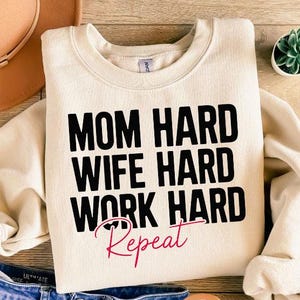May include: Cream-colored sweatshirt with the text "MOM HARD WIFE HARD WORK HARD Repeat" in black and pink. The sweatshirt is paired with blue jeans, a brown hat, sandals, and a small succulent plant.