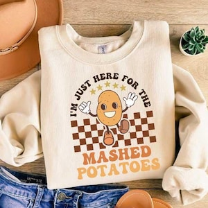May include: A beige sweatshirt with a brown and white checkered design and a cartoon potato character. The text on the sweatshirt reads "I'm just here for the mashed potatoes".