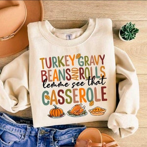 May include: A cream-colored sweatshirt with the text "TURKEY GRAVY BEANS AND ROLLS lemme see that CASSEROLE" in colorful letters. The sweatshirt has a fall-themed design with images of a pumpkin, a turkey, and a pie.