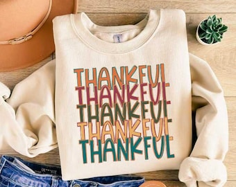 May include: Cream-colored sweatshirt with the word "THANKFUL" stacked multiple times in colorful, retro-style lettering. The image includes a brown hat, blue jeans, and sandals, suggesting a fall or Thanksgiving theme.