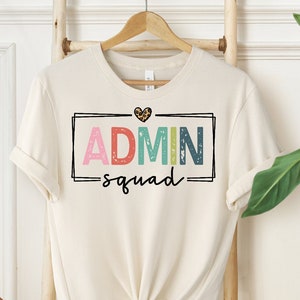 May include: A white t-shirt with a graphic design that says "ADMIN squad" in colorful letters with a small leopard print heart above the text.