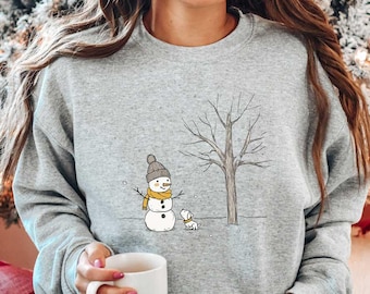 May include: A gray sweatshirt with a snowman and a dog illustration. The snowman is wearing a yellow scarf and a black hat. The dog is brown and white. The sweatshirt is being worn by a person sitting on a couch with a red and white checkered pillow.