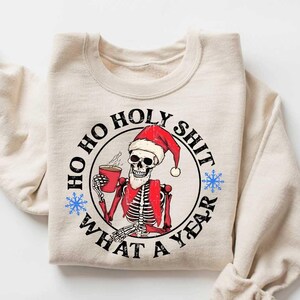 May include: A beige sweatshirt with a black and white graphic of a skeleton wearing a Santa hat and holding a cup of coffee. The graphic says "Ho Ho Holy Shit What A Year".
