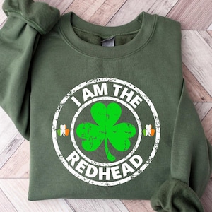 Puede incluir: Una sudadera verde con el texto "I am the redhead" en un c&iacute;rculo blanco con un tr&eacute;bol verde en su interior. El tr&eacute;bol tiene un contorno blanco y un centro verde.