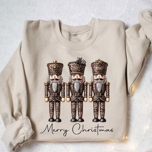 May include: A beige sweatshirt with a graphic of three nutcracker figurines wearing leopard print outfits. The text "Merry Christmas" is printed below the figurines.