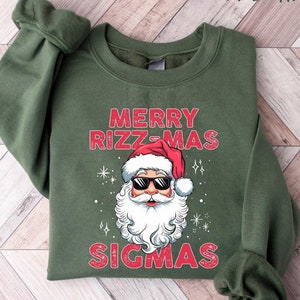 May include: Military green sweatshirt with the text "MERRY RIZZ-MAS SIGMAS" in red. The graphic features Santa Claus wearing sunglasses and a Santa hat. The sweatshirt is displayed on a light wood grain surface.