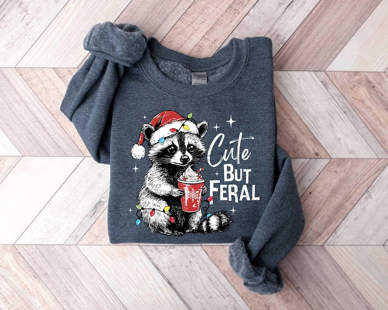 May include: A dark gray sweatshirt with a cartoon raccoon wearing a Santa hat and holding a red cup. The raccoon is surrounded by Christmas lights. The text "Cute But Feral" is printed on the sweatshirt.