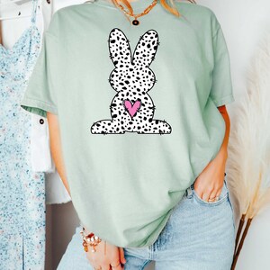 Pu&ograve; includere: Una maglietta verde chiaro con un coniglio macchiato di bianco e nero che tiene un cuore rosa. Il coniglio &egrave; un'illustrazione in stile cartone animato.