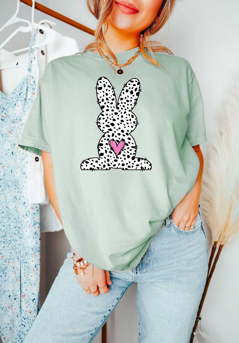 Pu&ograve; includere: Una maglietta verde chiaro con un coniglio macchiato di bianco e nero che tiene un cuore rosa. Il coniglio &egrave; un'illustrazione in stile cartone animato.