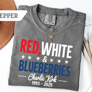 May include: A gray t-shirt with the words "RED, WHITE & BLUEBERRIES" in red, white, and blue. The text "Charlie Kirk 1993-2025" is below. A silver necklace and a brown hat are also visible.