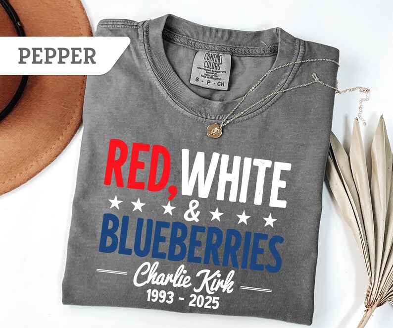 May include: A gray t-shirt with the words "RED, WHITE & BLUEBERRIES" in red, white, and blue. The text "Charlie Kirk 1993-2025" is below. A silver necklace and a brown hat are also visible.