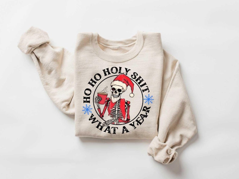 May include: A beige sweatshirt with a black and white graphic of a skeleton wearing a Santa hat and holding a cup of coffee. The graphic says "Ho Ho Holy Shit What A Year".