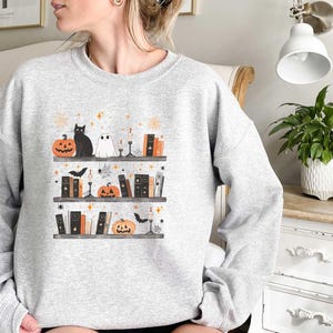 May include: A gray sweatshirt with a Halloween-themed design featuring a black cat, a ghost, and a bookshelf filled with books and pumpkins. The design is in a cartoon style and includes stars and other decorative elements.