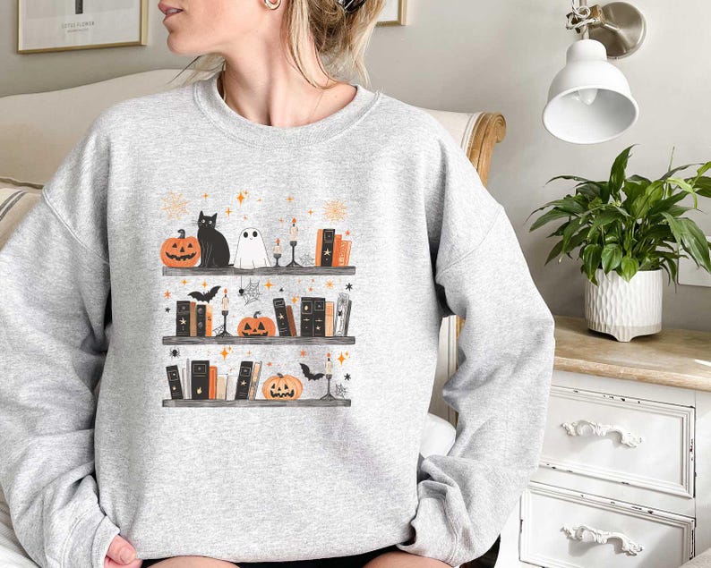 May include: A gray sweatshirt with a Halloween-themed design featuring a black cat, a ghost, and a bookshelf filled with books and pumpkins. The design is in a cartoon style and includes stars and other decorative elements.