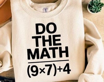 May include: Cream-colored sweatshirt with the text "DO THE MATH" in bold black letters. Below the text is the equation (9x7)+4. The sweatshirt is folded and placed on a wooden surface.