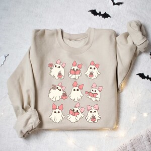 May include: Beige sweatshirt featuring a repeating pattern of cartoon ghosts with pink bows. Some ghosts hold flowers, books, or baskets. Black bat cutouts and string lights decorate the background, creating a Halloween theme.