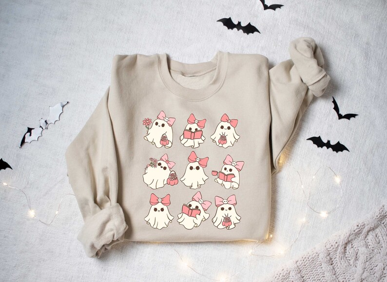 May include: Beige sweatshirt featuring a repeating pattern of cartoon ghosts with pink bows. Some ghosts hold flowers, books, or baskets. Black bat cutouts and string lights decorate the background, creating a Halloween theme.