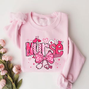 Peut inclure: Sweat-shirt rose avec un motif graphique du mot "Nurse" en rose et blanc avec des c&oelig;urs, un n&oelig;ud et des symboles m&eacute;dicaux.