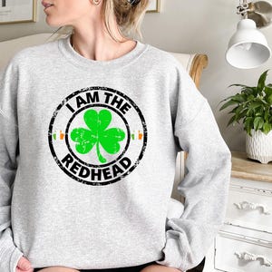 Puede incluir: Una sudadera gris con un dise&ntilde;o circular negro que dice "I am the Redhead" con un tr&eacute;bol verde dentro del c&iacute;rculo.
