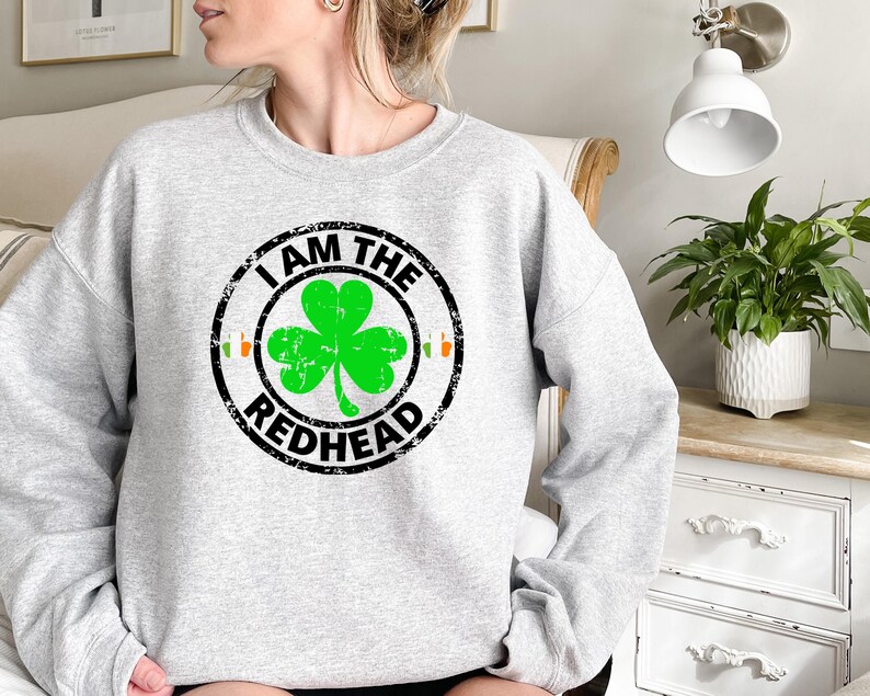 Puede incluir: Una sudadera gris con un dise&ntilde;o circular negro que dice "I am the Redhead" con un tr&eacute;bol verde dentro del c&iacute;rculo.