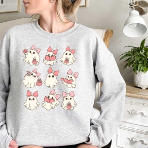 May include: A light gray sweatshirt featuring a repeating pattern of cartoon ghosts. Each ghost has a pink bow and is holding various items, including flowers, books, and baskets. The design is whimsical and cute.