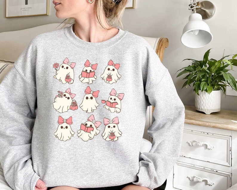 May include: A light gray sweatshirt featuring a repeating pattern of cartoon ghosts. Each ghost has a pink bow and is holding various items, including flowers, books, and baskets. The design is whimsical and cute.