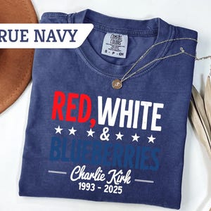 May include: A true navy blue t-shirt with the words "RED, WHITE & BLUEBERRIES" in red, white, and blue. The text "Charlie Kirk 1993-2025" is below the main text. A gold necklace is draped across the shirt.